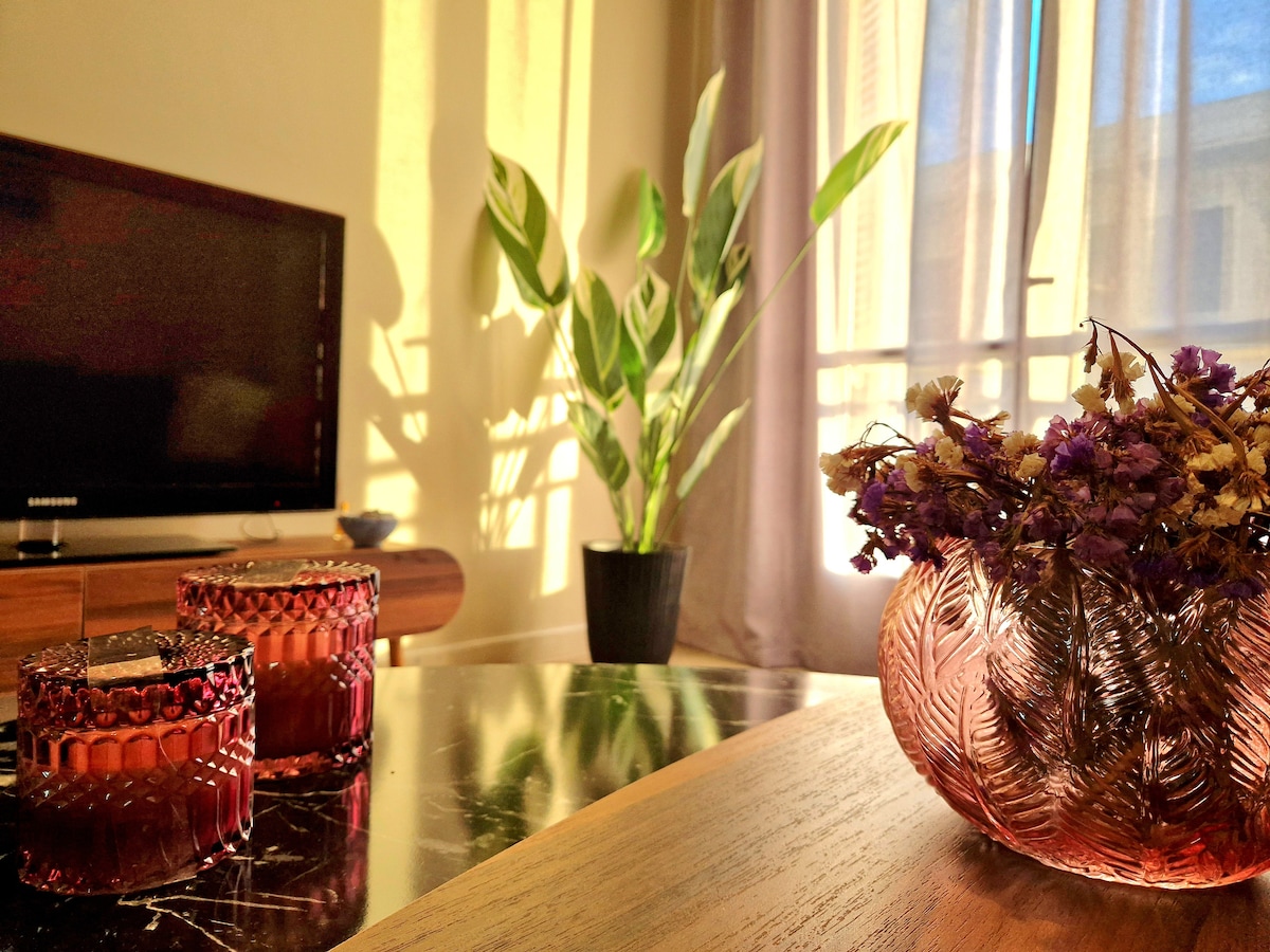 A cozy living area is illuminated by natural light filtering through sheer curtains. A tall plant stands in a black pot beside a wooden table. Two decorative candles and a glass vase filled with dried flowers add warmth to the space.