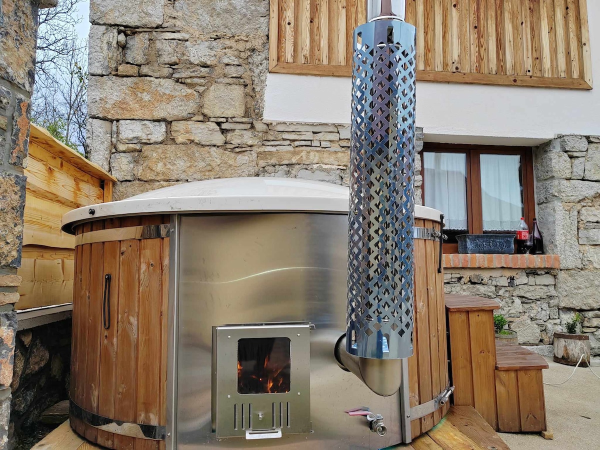 A wood-fired hot tub, constructed from wood and metal, is featured in a tranquil outdoor setting. The surrounding wooden decking complements the rustic stone walls of the house. A chimney extends upward, indicating the heating source, while a small fire can be seen inside the tub.