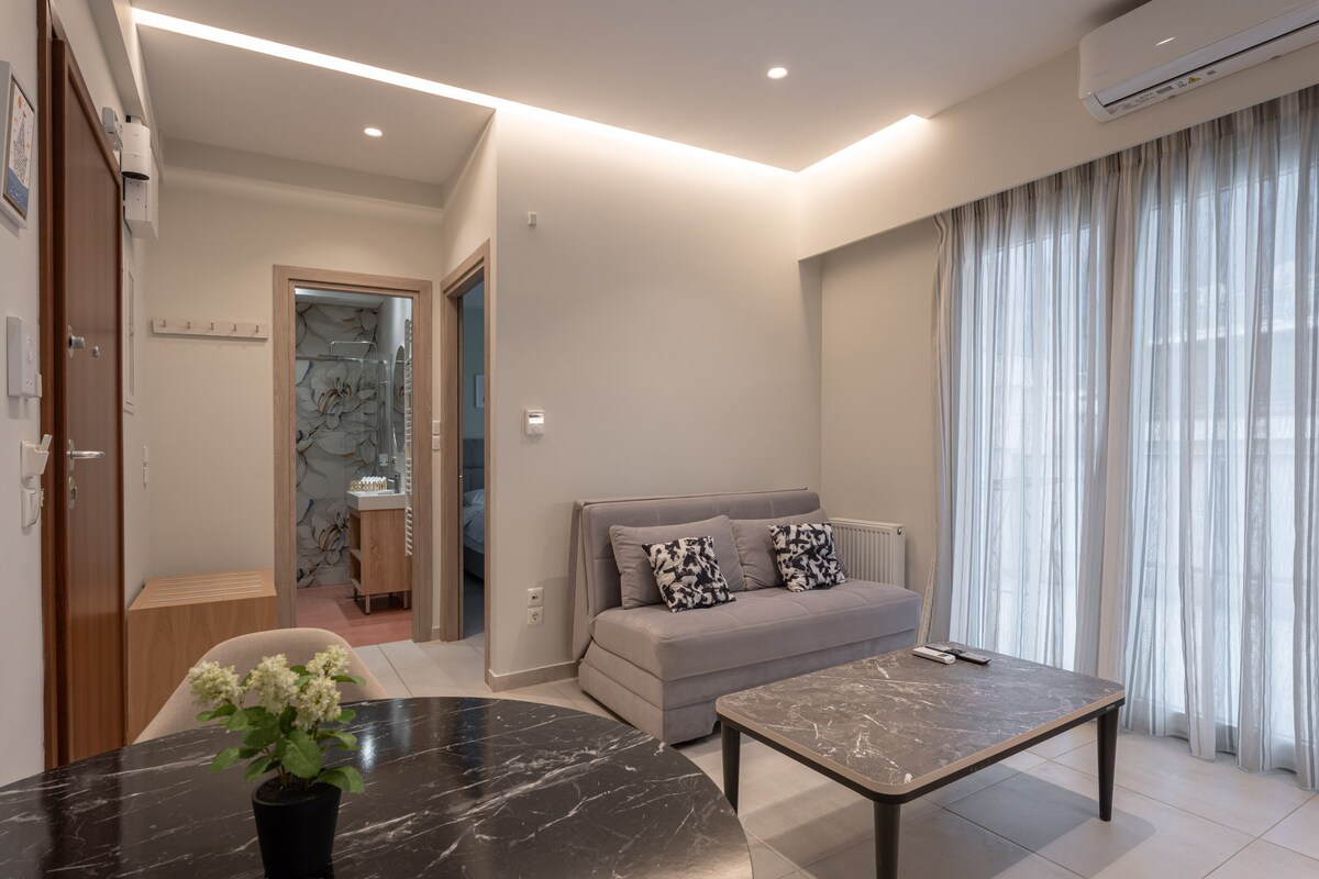 A well-lit living area is featured, showcasing a gray sofa adorned with decorative cushions. A sleek coffee table sits at the center, accompanied by a vase of flowers. Floor-to-ceiling windows allow natural light to fill the room, with soft curtains partially drawn.