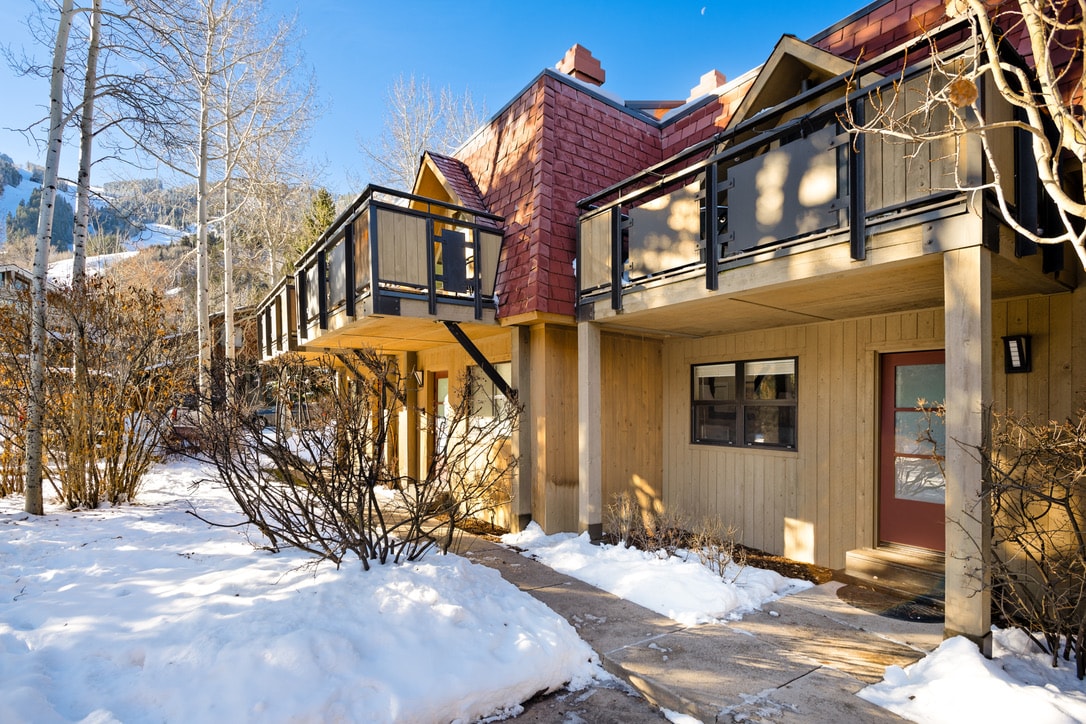 The exterior of a modern townhouse is visible, showcasing a combination of wood and red paneling. A private balcony extends over the entrance, surrounded by snow-covered landscaping and aspen trees. The clear sky suggests a sunny day, enhancing the mountain views in the background.