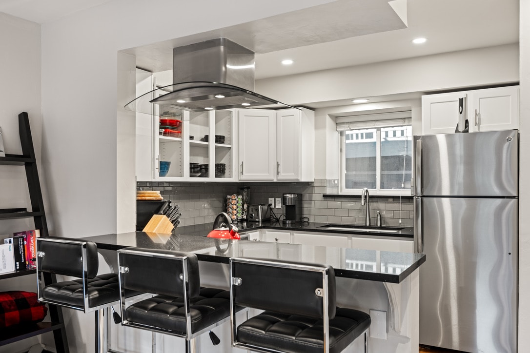 The modern kitchen features sleek white cabinetry with glass accents, a stainless steel refrigerator, and a gas stove. A polished gray countertop provides ample space for meal preparation, while three black bar stools are positioned at the kitchen island, creating a comfortable gathering spot.