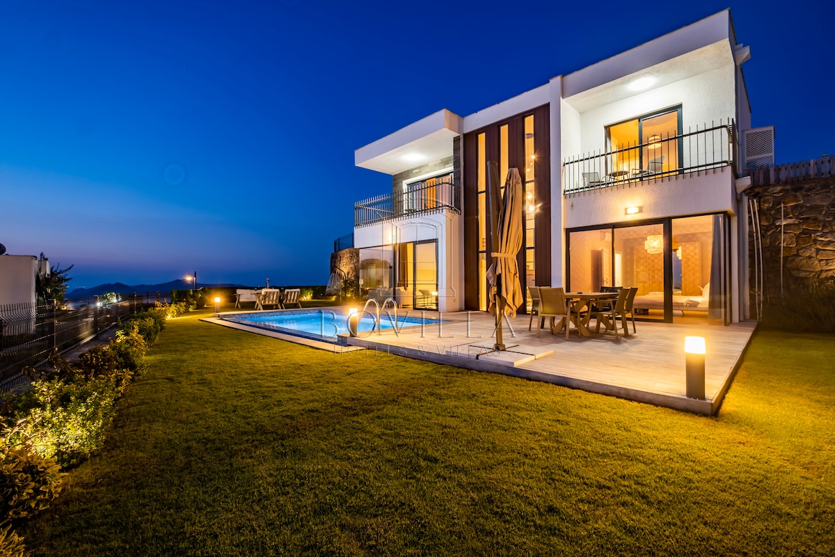 A modern villa is showcased against a twilight sky, with outdoor seating and a dining area visible near a sparkling swimming pool. Soft lighting illuminates the property, which is surrounded by a well-maintained lawn and includes a sun umbrella for added comfort.