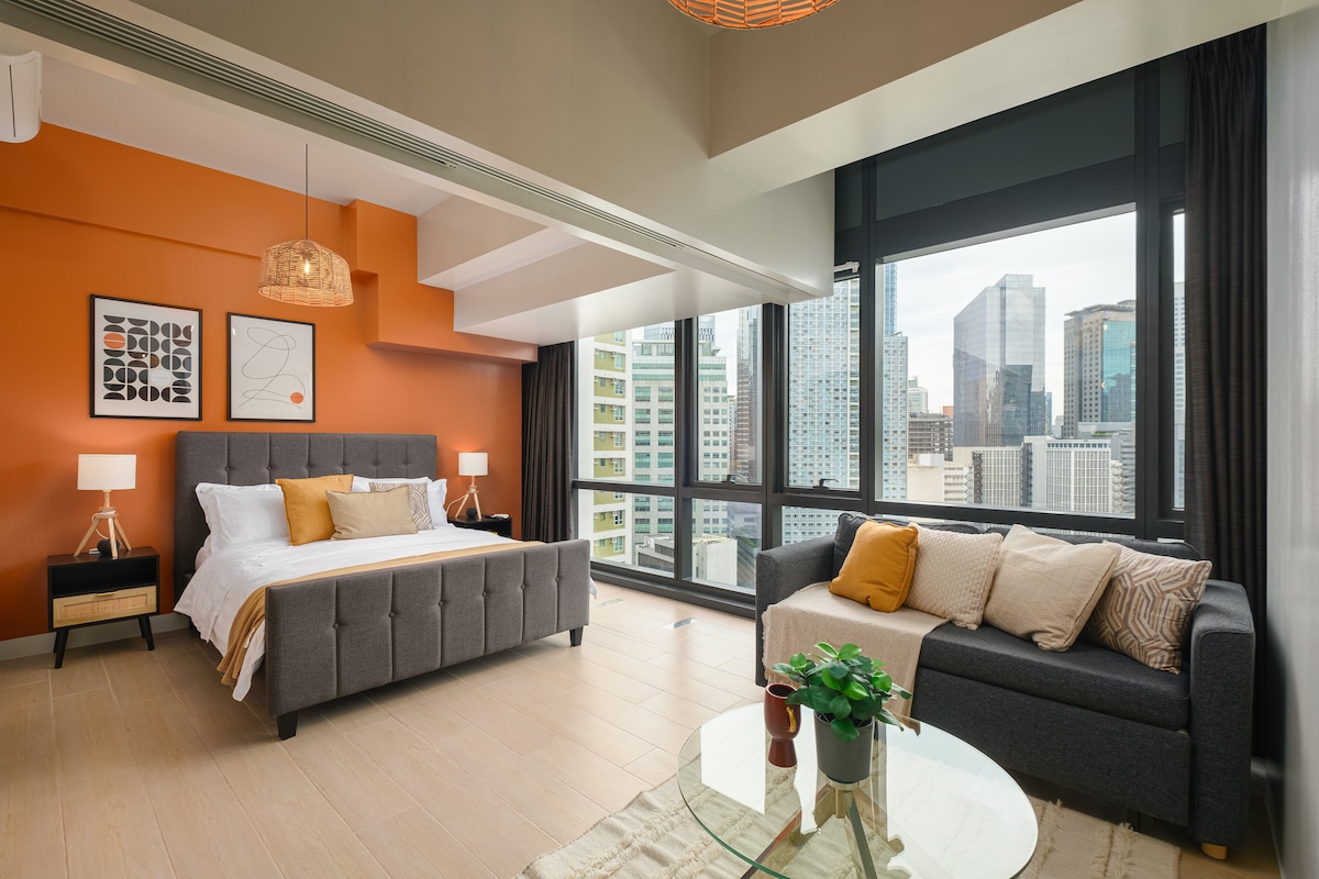 The bedroom features a king-sized bed dressed in soft linens, positioned near large windows showcasing city skyline views. A cozy sofa is placed adjacent, with a decorative coffee table and a potted plant completing the inviting space.