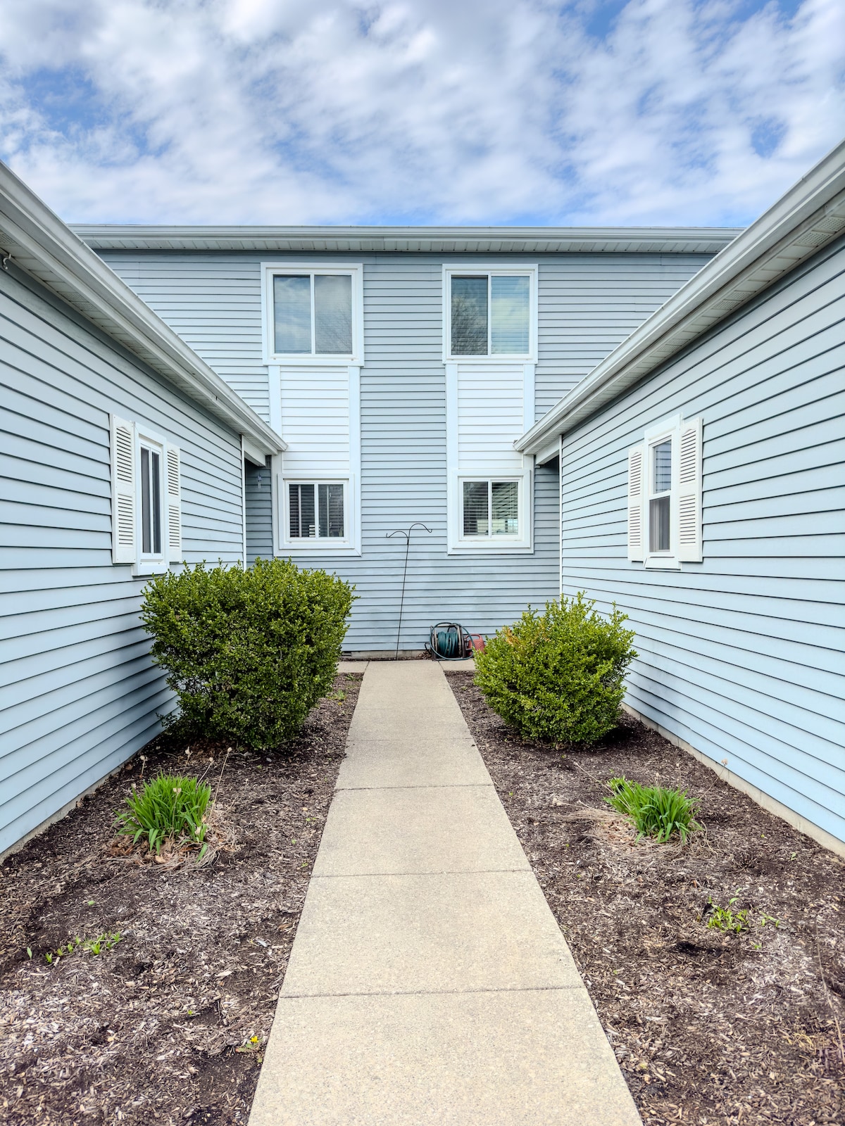 A well-maintained pathway leads to a two-story condominium, framed by landscaped areas featuring low shrubs and a flower bed. Light clouds are visible in the sky, creating a bright and inviting entrance.