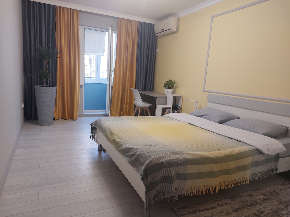 The spacious bedroom features a large bed with a soft blanket, accompanied by a desk and chair for work. Tall curtains frame the window, allowing natural light to fill the room. A potted plant adds a touch of greenery to the modern ambiance.