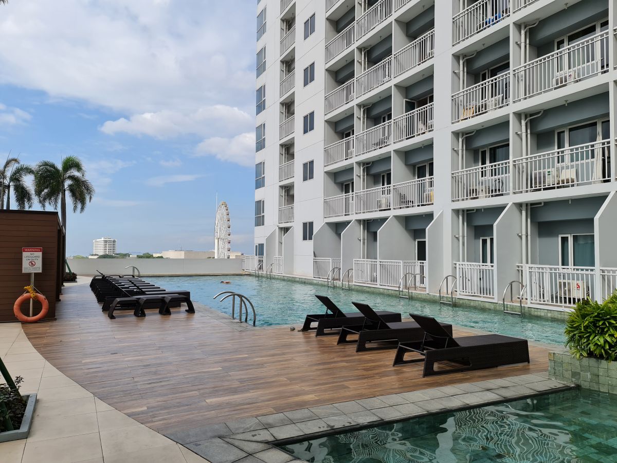 A serene swimming pool area is featured, surrounded by sleek wooden decking. Numerous lounge chairs are arranged for relaxation, with the modern condominium building visible in the background. A clear sky and water enhance the tranquil atmosphere of the space.