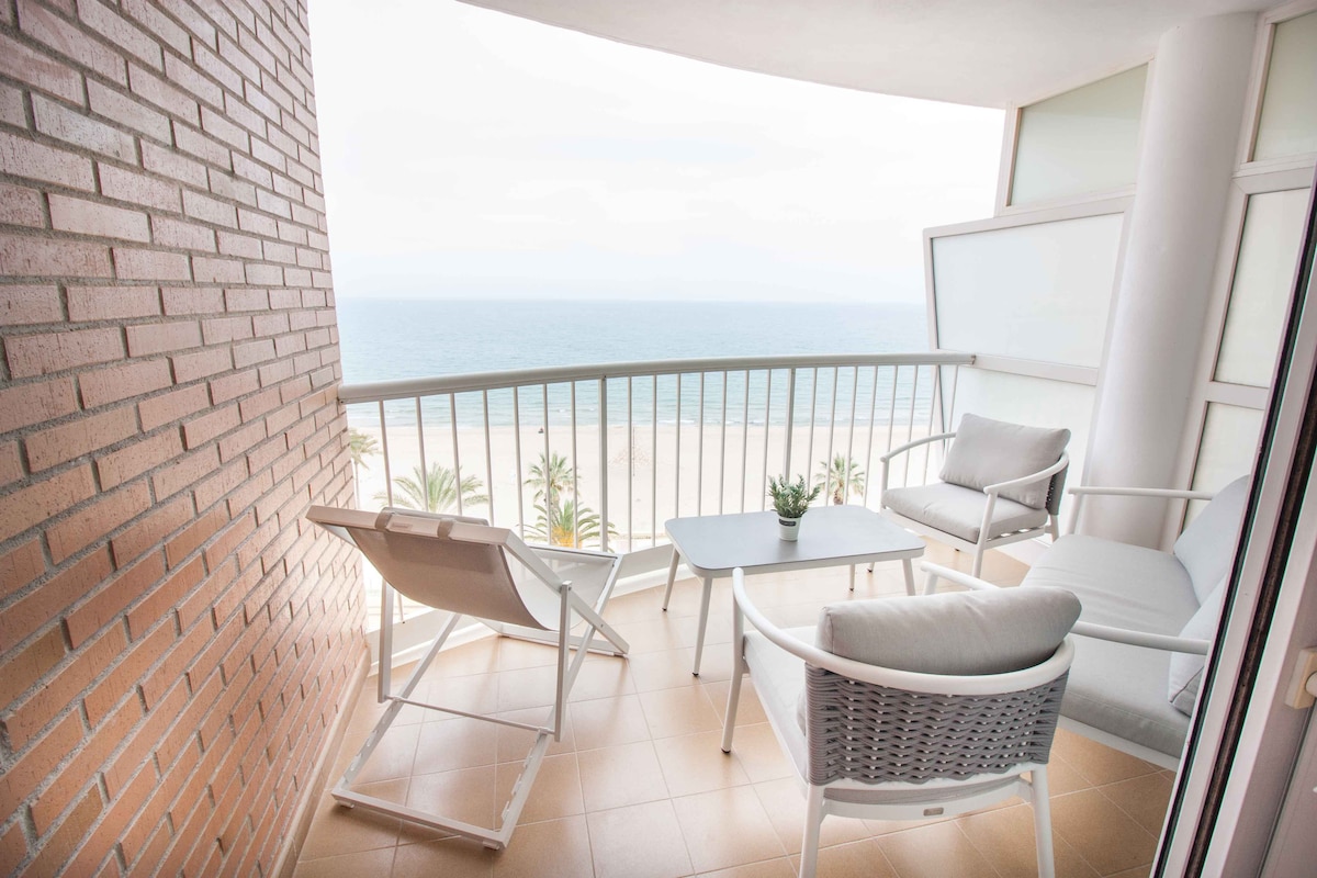 A balcony space features a table and chairs, allowing for views of the sea and coastline. The floor is tiled, and a small plant adorns the table. Sunlight can be seen illuminating the area, with railing providing safety and an enclosed feeling.