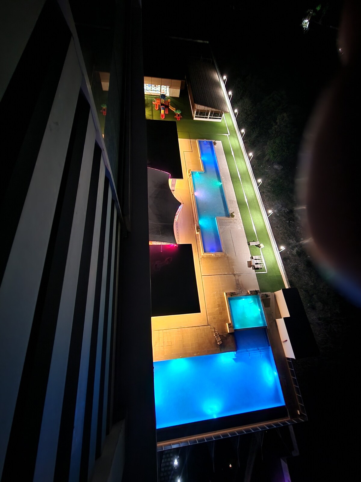 An aerial view of the pool area displays multiple swimming pools illuminated in vibrant shades of blue and purple. Surrounding amenities include lounging areas and pathways lined with lights, enhancing the tranquil nighttime atmosphere of the Royal Palms.