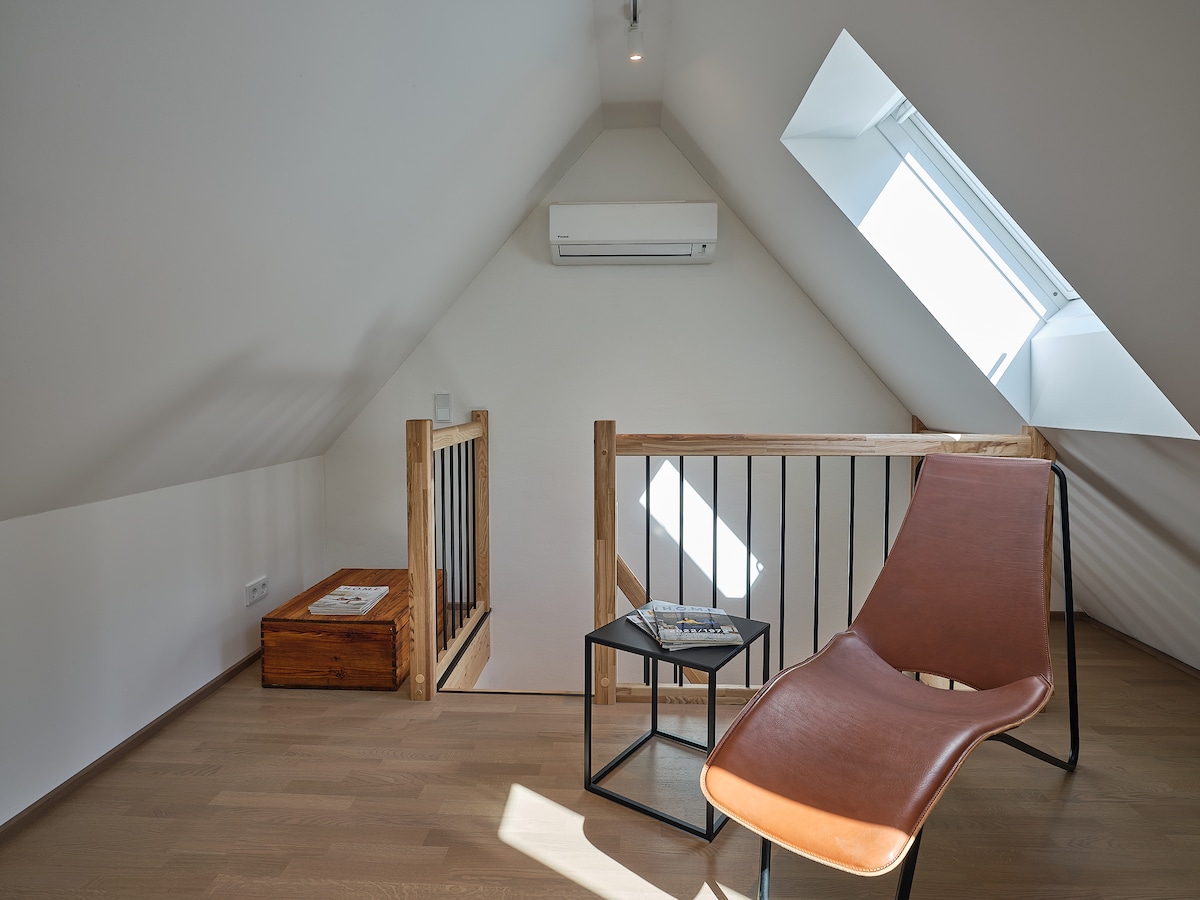 A cozy loft area is captured, showcasing a sleek, modern chaise lounge positioned next to a small black side table. Natural light streams through a skylight, illuminating the wooden floors and the minimalist wooden railings leading to a stairway.