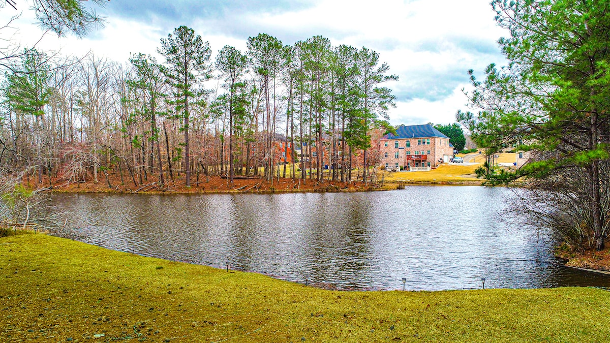 A serene lake is bordered by a grassy area and framed by tall trees in the background. Buildings are visible on the opposite bank, contributing to the tranquil landscape.