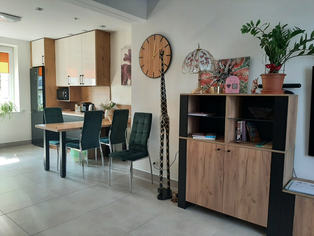 A modern kitchen and dining area is showcased, featuring a wooden dining table with six green upholstered chairs. A decorative giraffe figure stands beside a dark wooden shelving unit containing books and a plant. Natural light enters through the windows, enhancing the space.