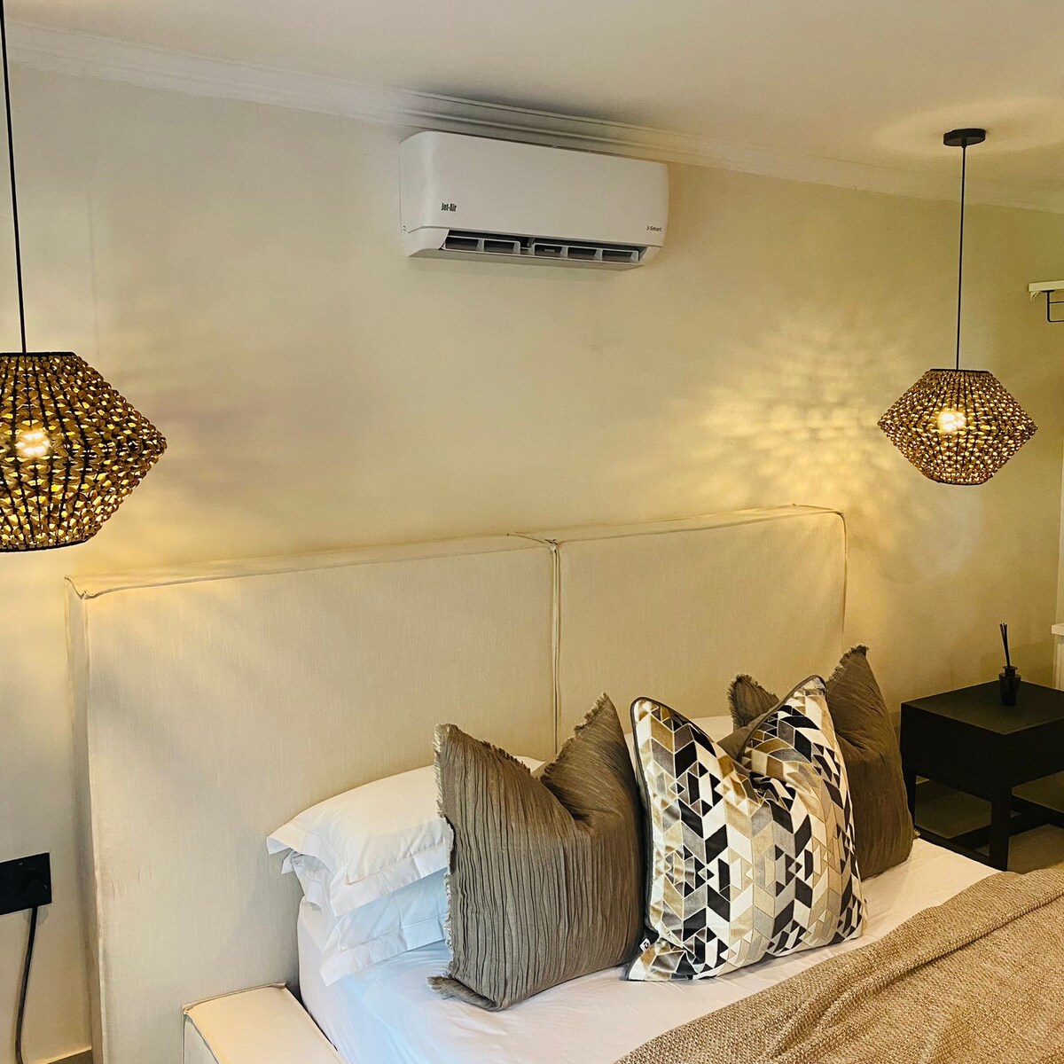 A sleeping area features a soft headboard adorned with decorative pillows in various patterns and textures. Two pendant lights provide a gentle glow from either side, enhancing the serene environment. An air conditioning unit is installed above, ensuring comfort throughout the space.