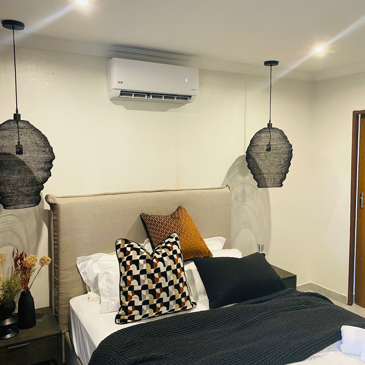 A modern bedroom displays a spacious bed with a neutral headboard. Two pendant lights hang above, with dark woven shades. Decorative pillows in various patterns and colors are arranged on the bed, while a small side table holds a vase with dried flowers.