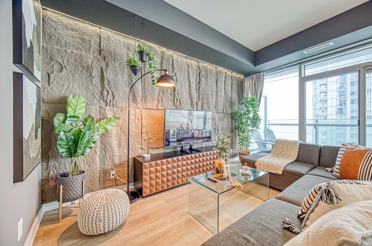 The living room features a modular sectional sofa arranged around a glass coffee table, complemented by a stone accent wall and greenery. A 55'' TV is mounted on the wall, with natural light pouring in through large windows that provide skyline views.
