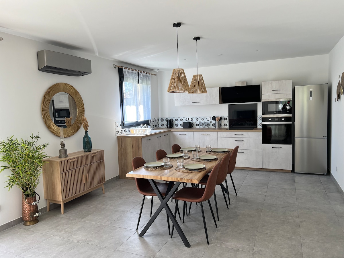 A modern kitchen and dining area features a wooden table set for six with elegant dishware. The kitchen includes contemporary appliances and cabinetry, complemented by decorative tile accents. Natural light illuminates the space through large windows, while a potted plant adds a touch of greenery.