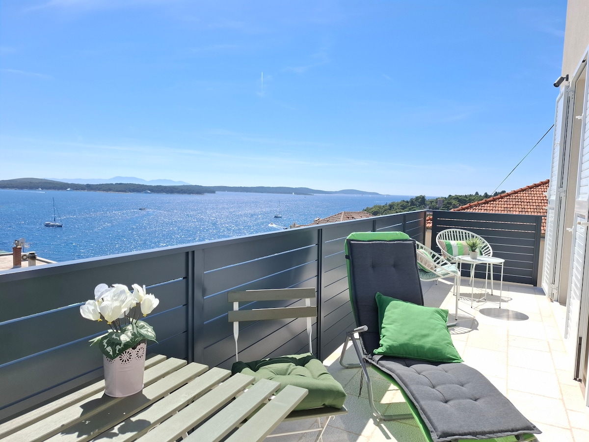 A spacious balcony offers stunning sea views, furnished with a green lounge chair and a matching cushion. A small table sits nearby, accompanied by two additional chairs. Decorative plants add a touch of greenery, enhancing the serene outdoor space.