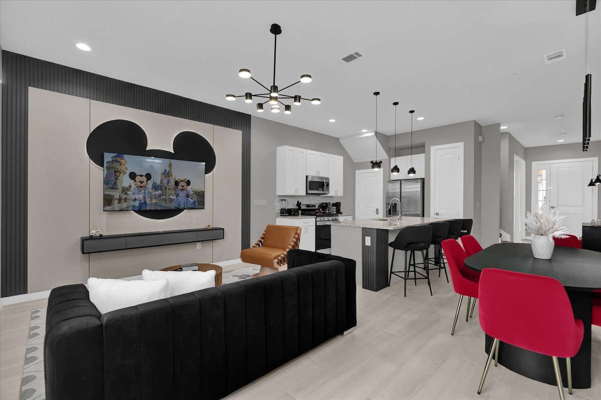 The family room is designed with a playful Mickey Mouse theme, showcasing a flat-panel TV mounted on a textured wall. A cozy black sectional sofa sits beside a tan armchair, and a modern dining table with red chairs occupies the adjacent space, creating an inviting atmosphere.