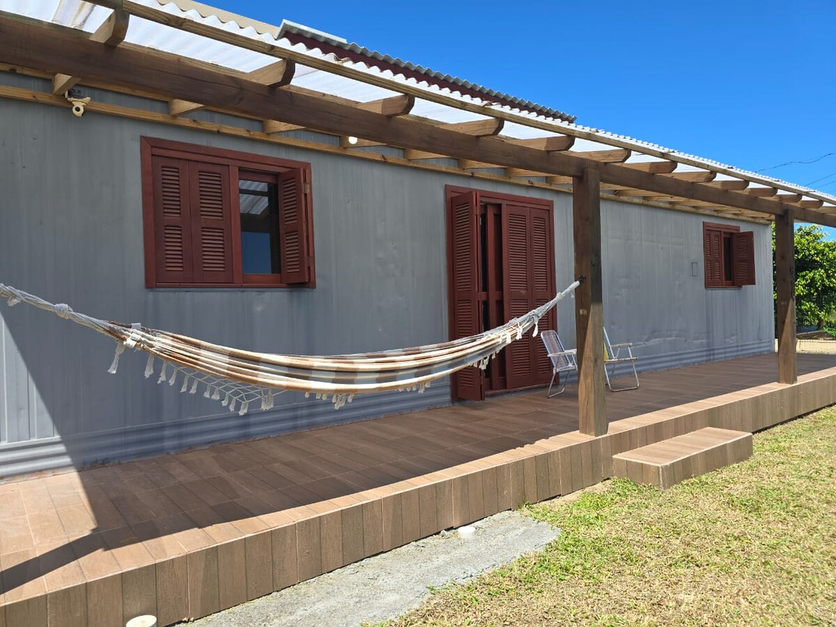 A relaxing porch is featured, showcasing a hammock suspended between wooden posts. Two chairs are placed nearby under a pergola, with red-shuttered windows adding to the charming facade. The surrounding grass complements the natural setting, bathed in bright daylight.
