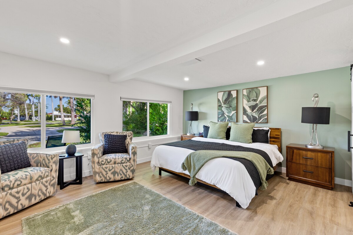 A spacious bedroom is highlighted by a large bed dressed in crisp linens and layered with a green throw blanket. Two chairs are positioned near large windows, allowing natural light to fill the room. Art featuring botanical designs adorns the wall, complementing the soft color palette.