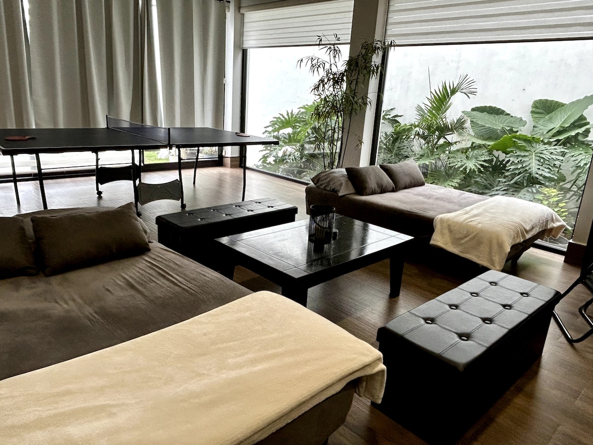 A spacious interior features two comfortable sofa beds set against large windows, allowing natural light to illuminate the room. A dark wood table sits in the center, complemented by a ping-pong table. Lush greenery is visible outside, enhancing the inviting atmosphere.