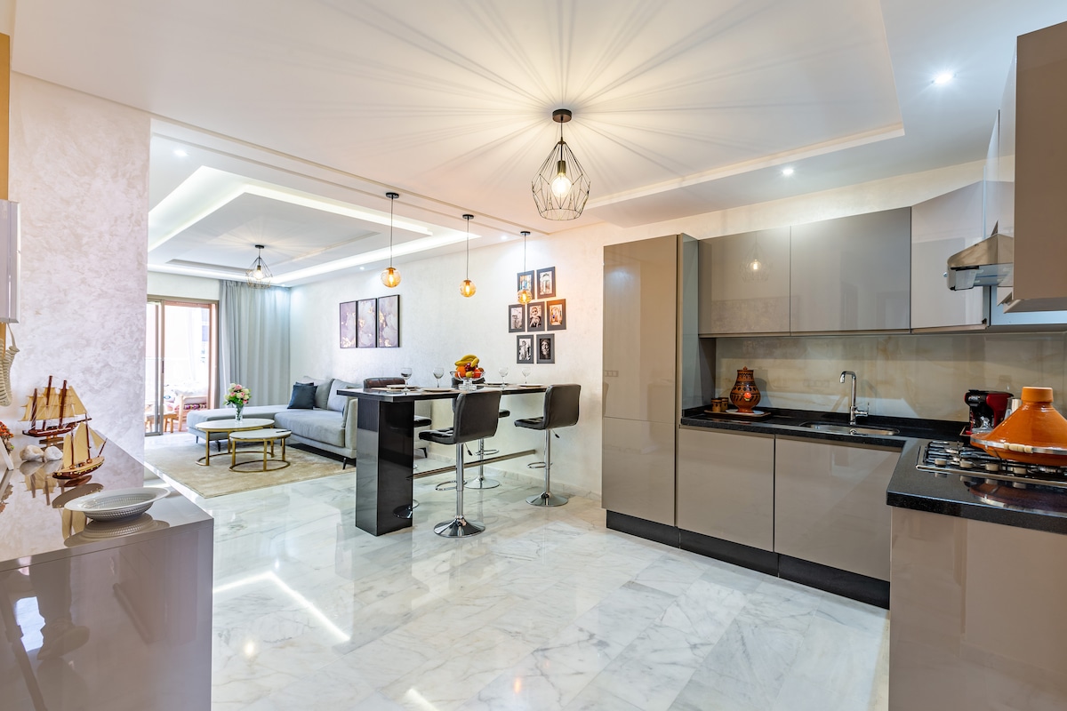An open-concept living area features modern furnishings with a spacious kitchen, a dining table and four chairs, and a relaxing seating area. Natural light floods in from large windows, accentuated by tasteful decor on the walls, creating a bright and inviting atmosphere.