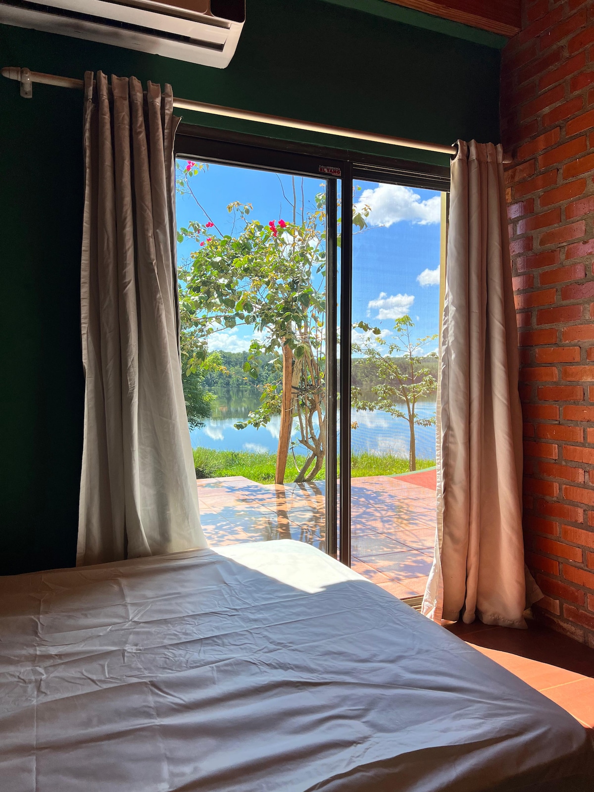A spacious bedroom features large glass doors that open to a serene view of Lake Urugua-í. Natural light floods the room, highlighting the brick wall and soft white bedding. Outside, the peaceful lake reflects the blue sky and surrounding greenery.