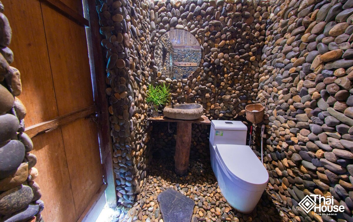A private bathroom is showcased, featuring walls and floor adorned with smooth river stones. A circular mirror hangs above a stone sink, while a wooden stand supports eco-toiletries. A toilet is situated adjacent, contributing to a natural and rustic atmosphere.