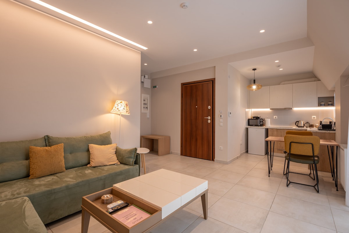 A modern living area features a green sofa with decorative cushions, positioned next to a sleek coffee table. A neutral wall creates a soft backdrop, while warm lighting enhances the space. An open kitchenette with dining chairs is visible, providing a functional area for meals.