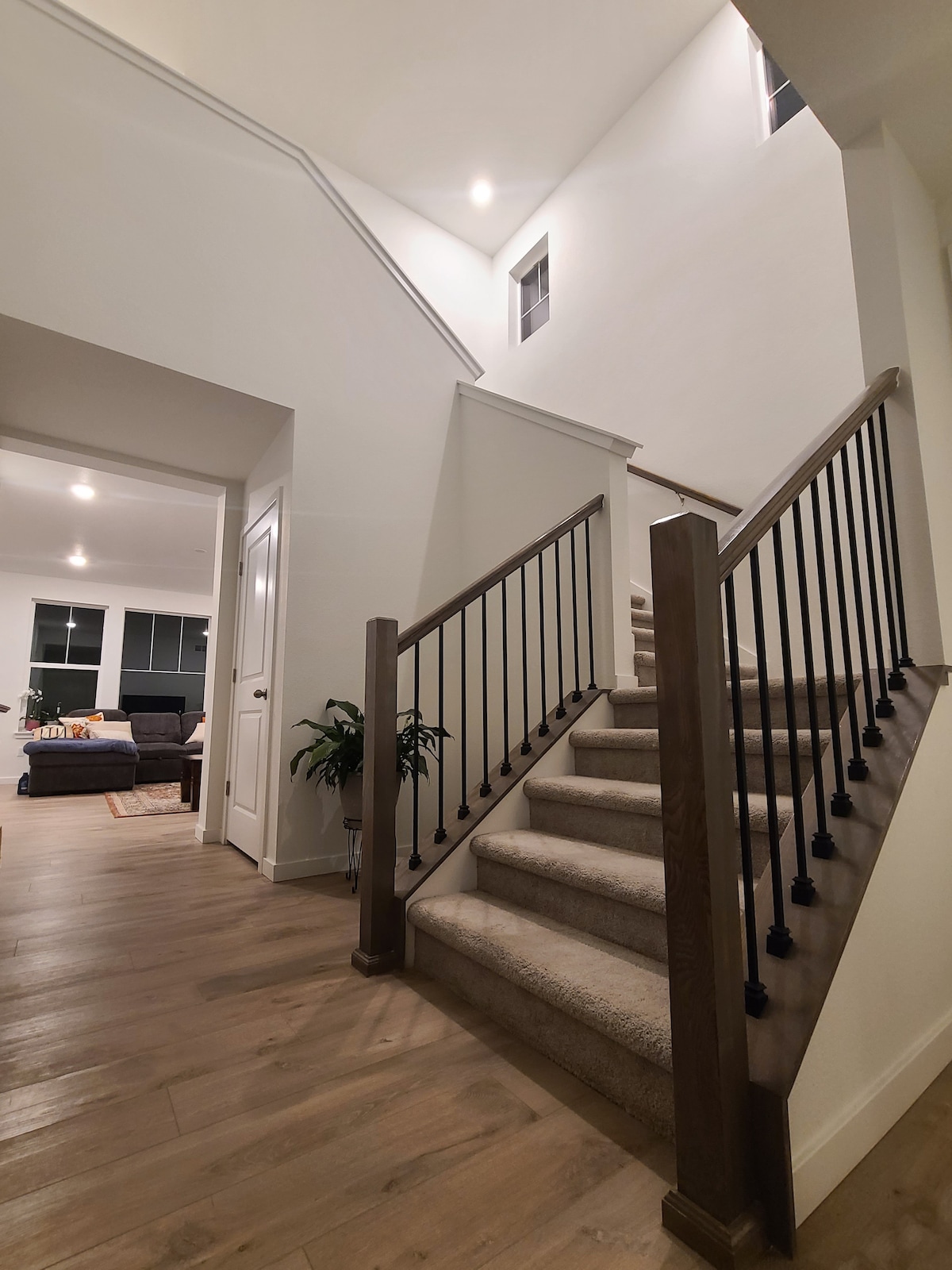 A well-lit staircase is seen leading to the second floor. The carpeted steps are framed by wooden railings with metal balusters. Light fixtures illuminate the area, highlighting white walls and warm-toned flooring. A glimpse of a cozy living area is visible in the background.
