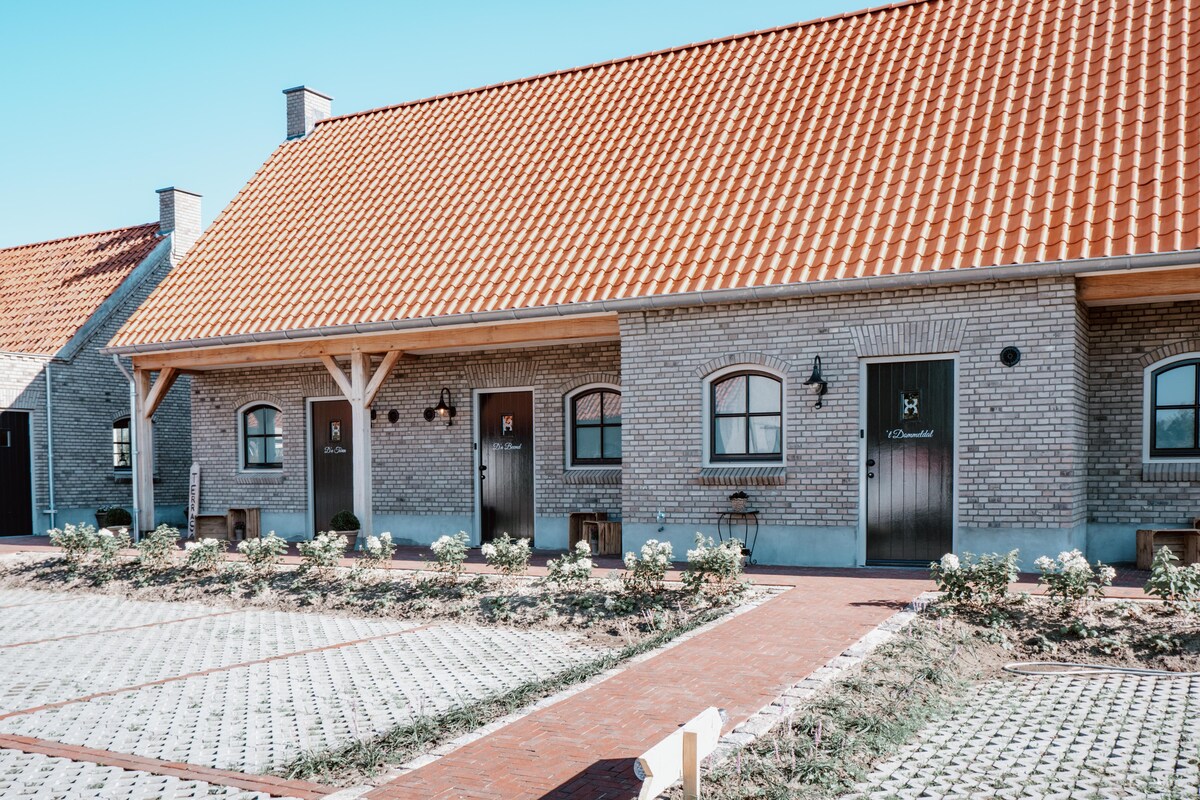 A row of modern brick cottages features welcoming wooden doors and windows, framed by neatly landscaped flower beds. The red-tiled roof adds a traditional touch, while a paved pathway leads to each entrance, enhancing accessibility. Ample parking is available in front of the cottages.