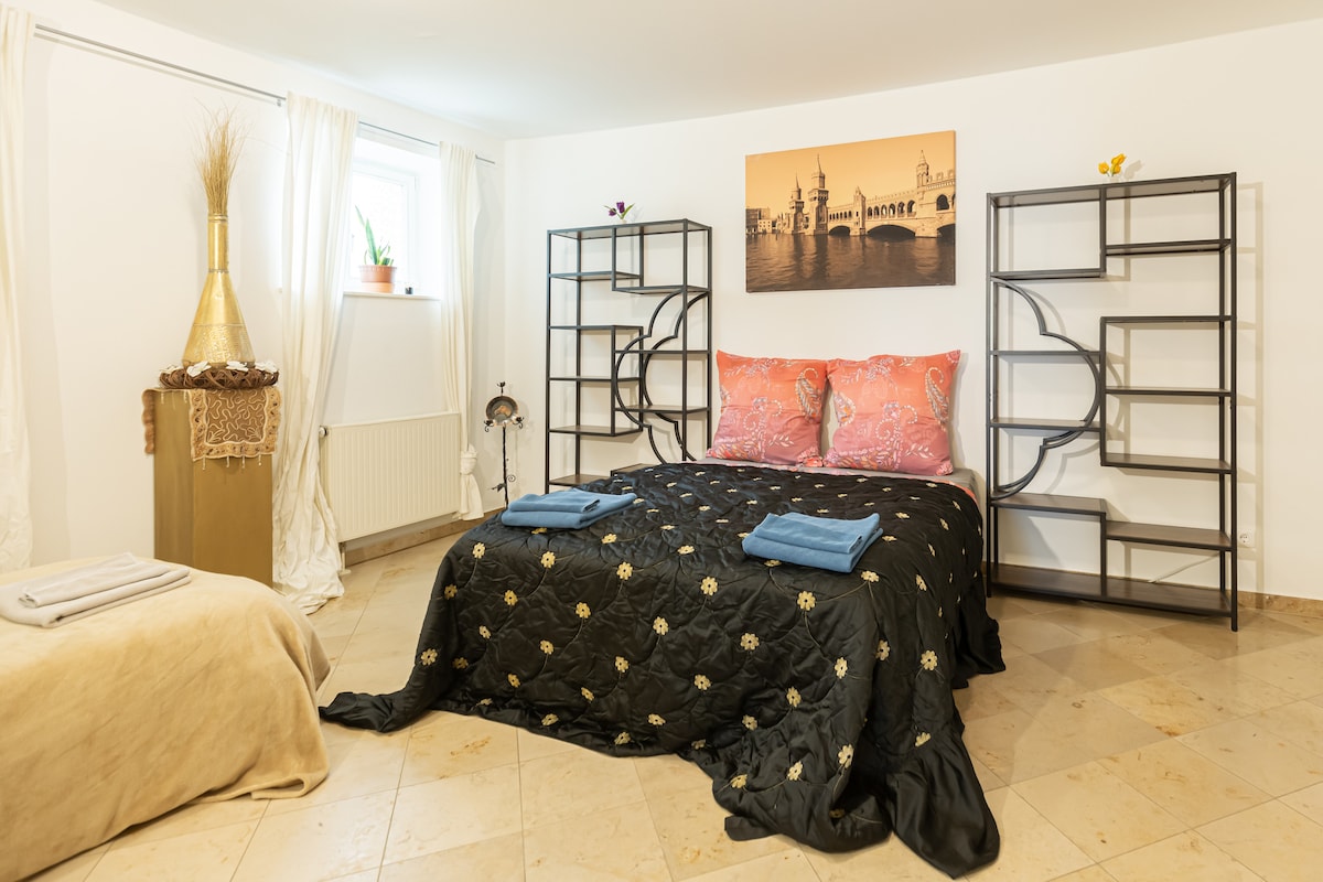 A sleeping area features a spacious bed dressed in a dark patterned coverlet, complemented by colorful pillows. Two open shelving units flank the bed, showcasing minimal decor. A window allows natural light to brighten the space, with sheer curtains draping on the sides.
