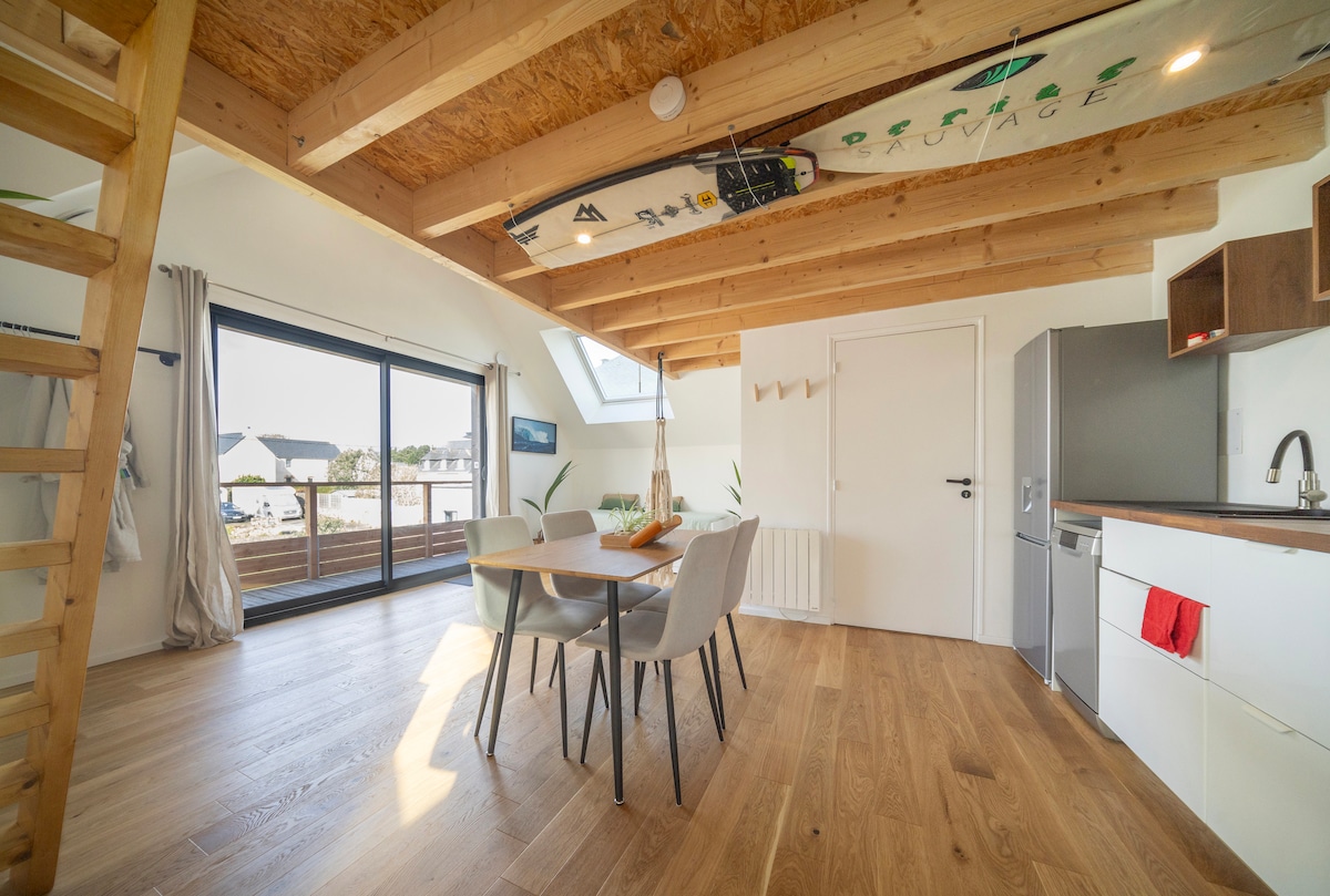 An open living area is highlighted by a wooden ceiling and large glass doors that invite natural light. A dining table for four is positioned next to the kitchen, which features modern appliances. A ladder leads to a mezzanine space, adding dimension to the layout.