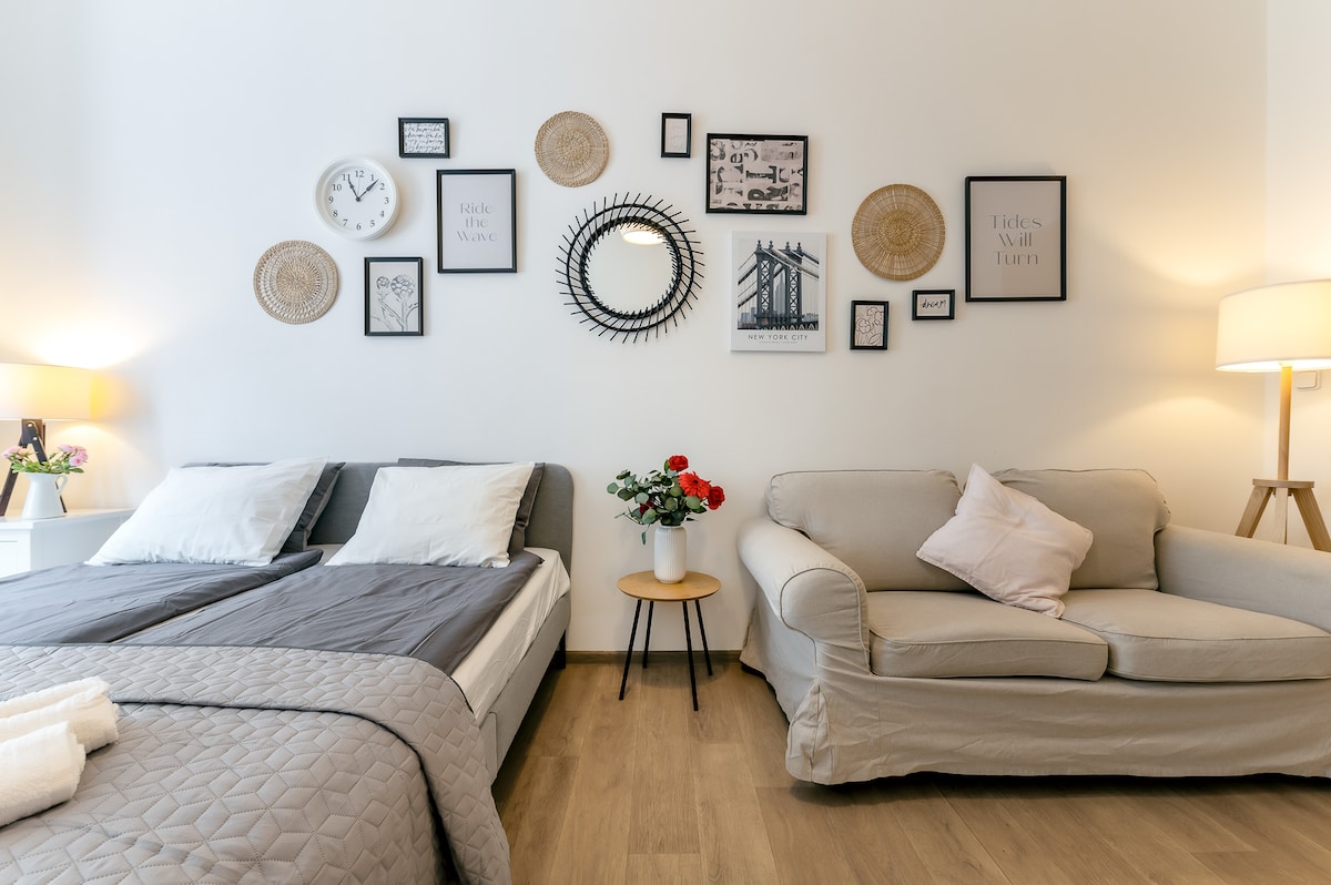 A cozy studio interior features a spacious bed made with light bedding beside a soft couch. Various decorative wall art, including clocks and framed prints, create an engaging focal point. A small round table holds a vase with fresh flowers, enhancing the inviting atmosphere.