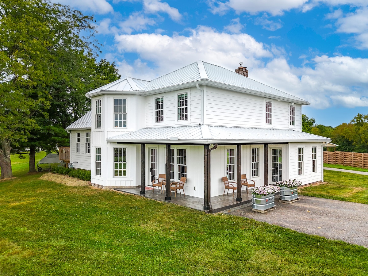 A two-story house features a classic white exterior, complemented by a covered porch. Large windows allow ample natural light, while comfortable outdoor seating is arranged on a patio. The surrounding grassy area is bordered by trees and a wooden fence, contributing to a peaceful setting.