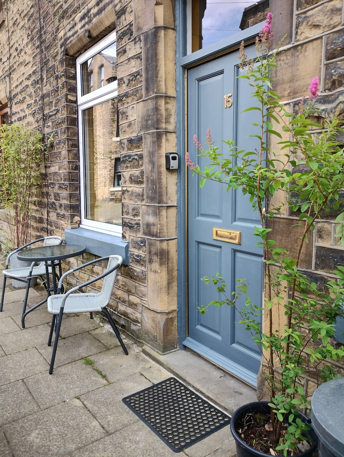 The entrance of the terraced house features a blue door with a gold handle, flanked by a stone facade. A small mat lies at the threshold, while two grey chairs and a round table are positioned on the cobblestone path, surrounded by green plants.