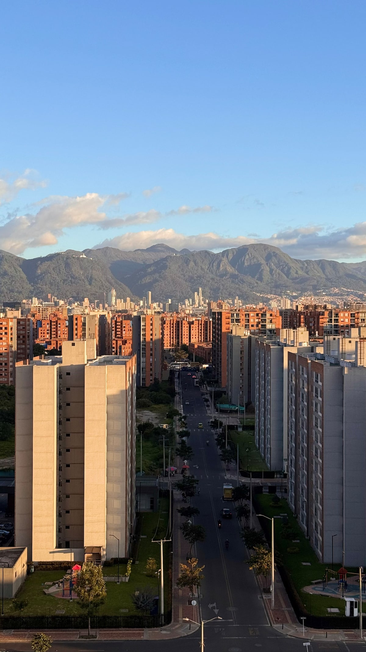 A panoramic view showcases modern residential buildings lining a wide avenue, extending towards a backdrop of rolling mountains. The scene is enriched with vibrant city life and greenery, illuminated under a clear blue sky that enhances the visual contrast.
