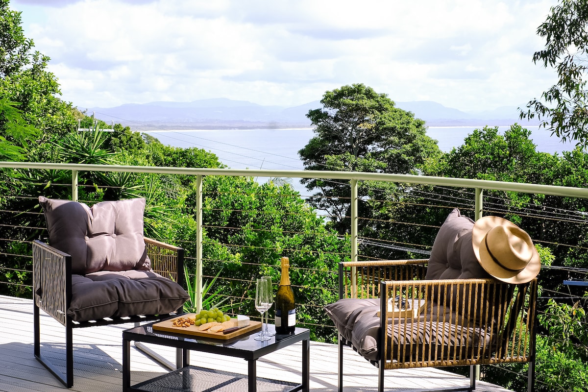 An outdoor lounge area is portrayed on the deck, featuring two cushioned chairs beside a low table. A bottle of champagne and glasses sit on the table, with scenic views of lush greenery and the bay in the background.