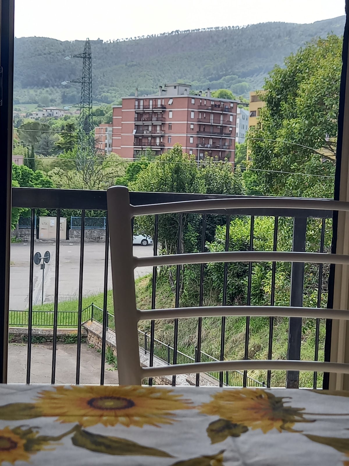 A view of the external surroundings is captured through a large window. A white chair partially frames the scene, which features lush greenery and a multi-story building in the distance. Natural light brightens the area, highlighting the urban and natural blend outside.