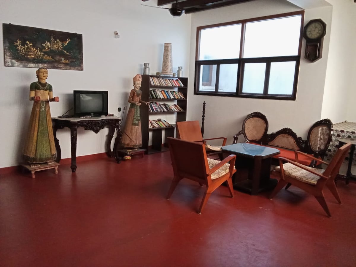 A spacious living room features a combination of traditional and contemporary decor. Two wooden chairs and a square table occupy the center. A vintage television and a bookshelf filled with various titles are visible. The walls are adorned with art pieces, and a decorative clock hangs in the corner.