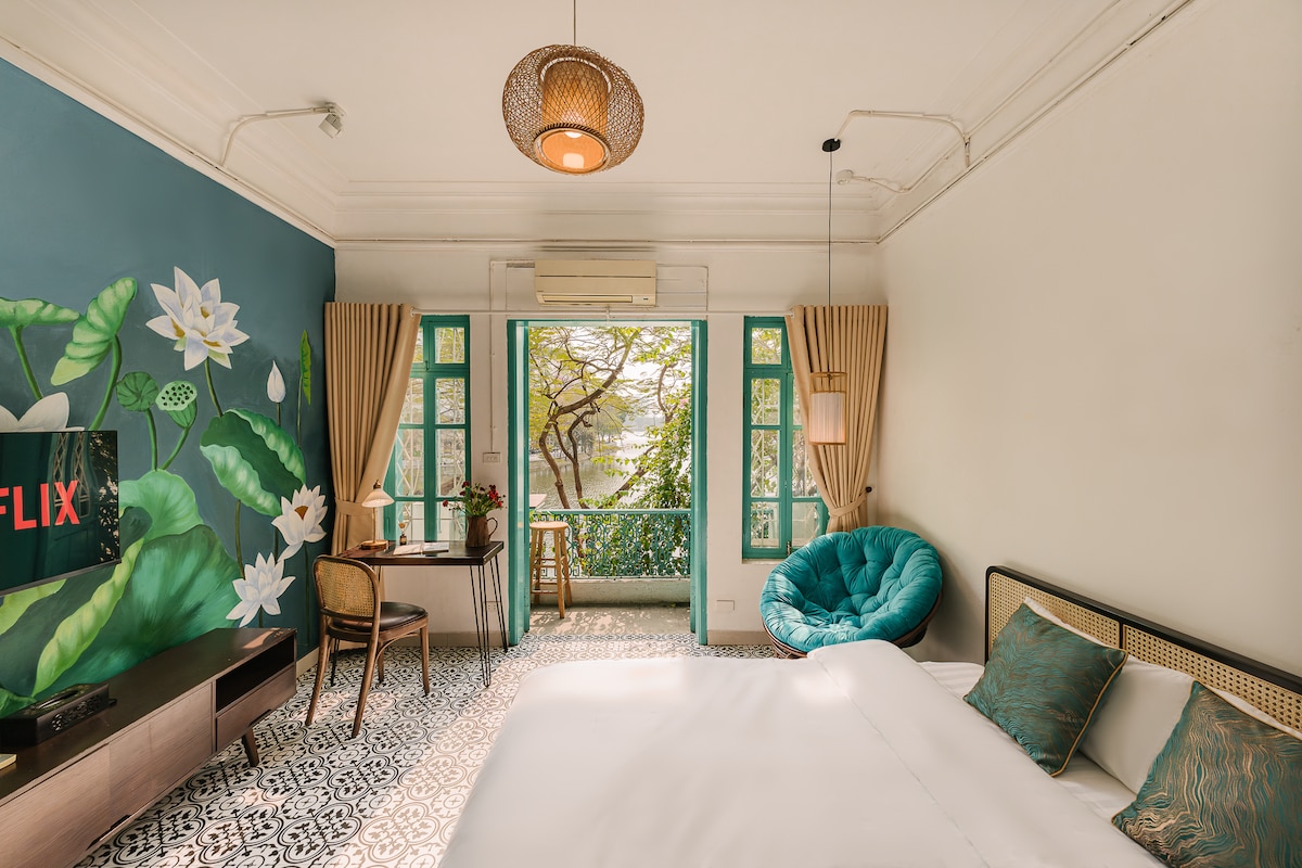 A serene room with a spacious bed and vibrant wall art featuring green foliage and lotus flowers. Large windows invite natural light and offer views of the outdoors. A cozy turquoise chair and a wooden desk create a functional workspace, complemented by soft drapes.