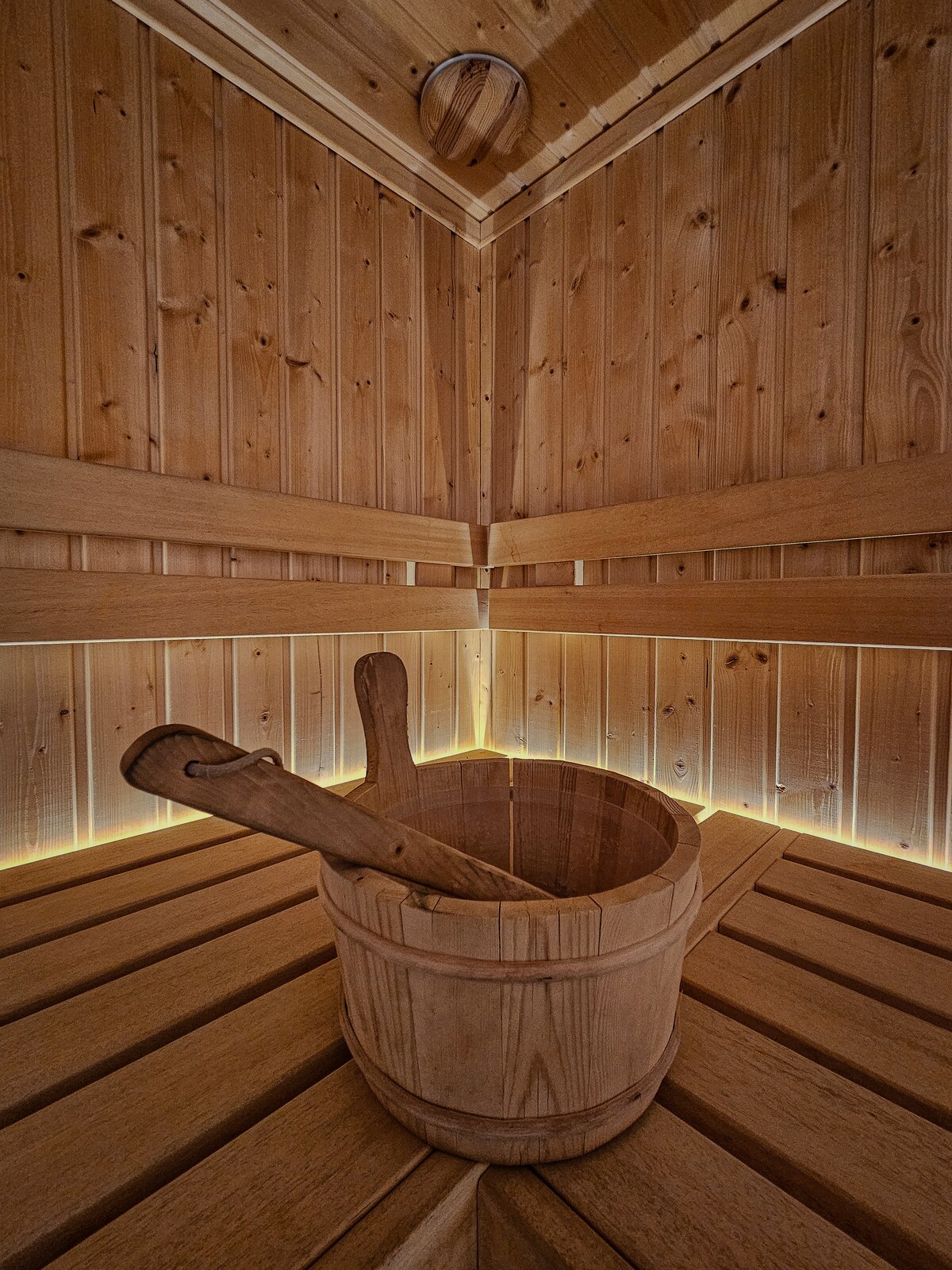 An inviting sauna space is shown, featuring warm wooden paneling and benches. Soft lighting creates a calming environment. A wooden bucket rests on the bench, suggesting traditional sauna use. The minimalist design focuses on natural materials, enhancing relaxation and wellness.