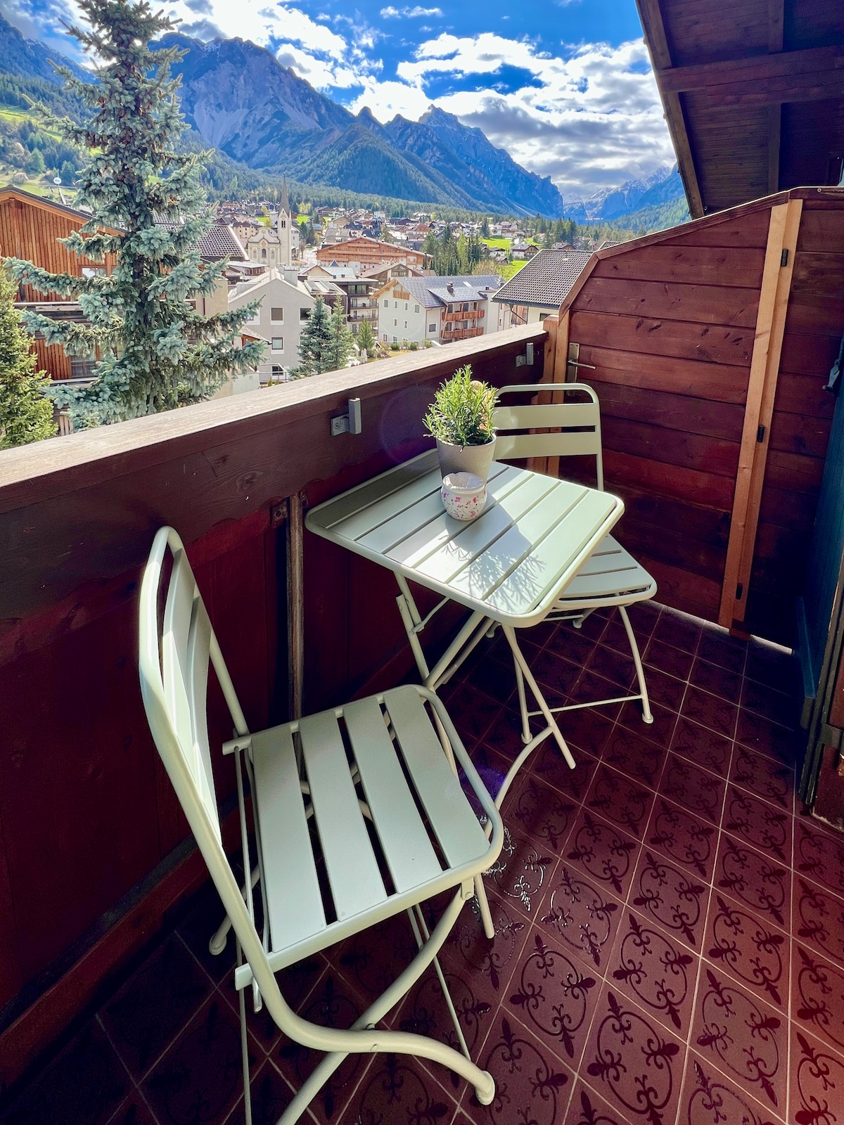 A small balcony is showcased, featuring a round table with a potted plant atop it and two chairs. Wooden structures and scenic mountain views are visible in the background, offering a serene outdoor space.