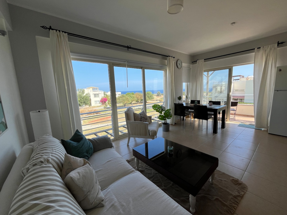 A bright living area presents large windows that frame a sea view. A comfortable sofa is positioned beside a coffee table, with a plant adding a touch of greenery. Dining space is organized with a table and chairs, allowing for dining with a view.