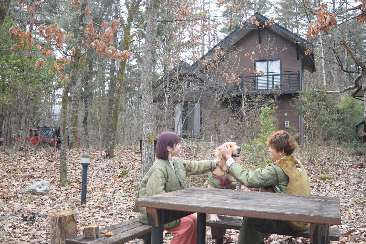 An outdoor setting features a rustic wooden table surrounded by fallen leaves, where two guests interact with a dog. The house is situated in the background among trees, showcasing a blend of natural surroundings and cozy cabin architecture.