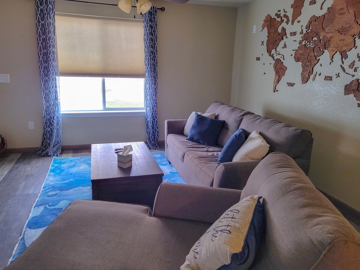 A comfortable living space features a large gray sectional sofa adorned with various textured cushions. A rectangular wooden coffee table sits in the center on a colorful blue rug. A large window with light-filtering shades allows natural light to brighten the room, complemented by a world map artwork on the wall.