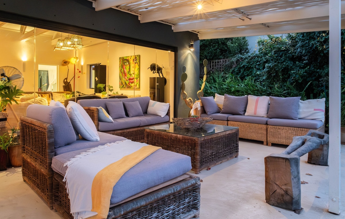 A spacious outdoor lounge area is featured, furnished with comfortable wicker seating adorned with soft cushions. A coffee table rests in the center, while ambient lighting creates a cozy atmosphere. Lush greenery surrounds the patio, enhancing the tranquil setting.
