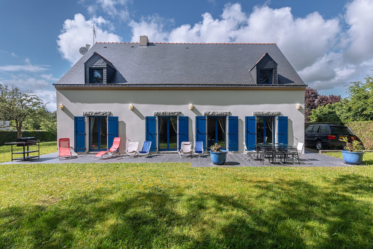 The exterior of the house features a two-story structure with large blue-shuttered windows and a gray roof. A spacious patio area includes loungers and a dining set surrounded by greenery, providing a comfortable outdoor space.