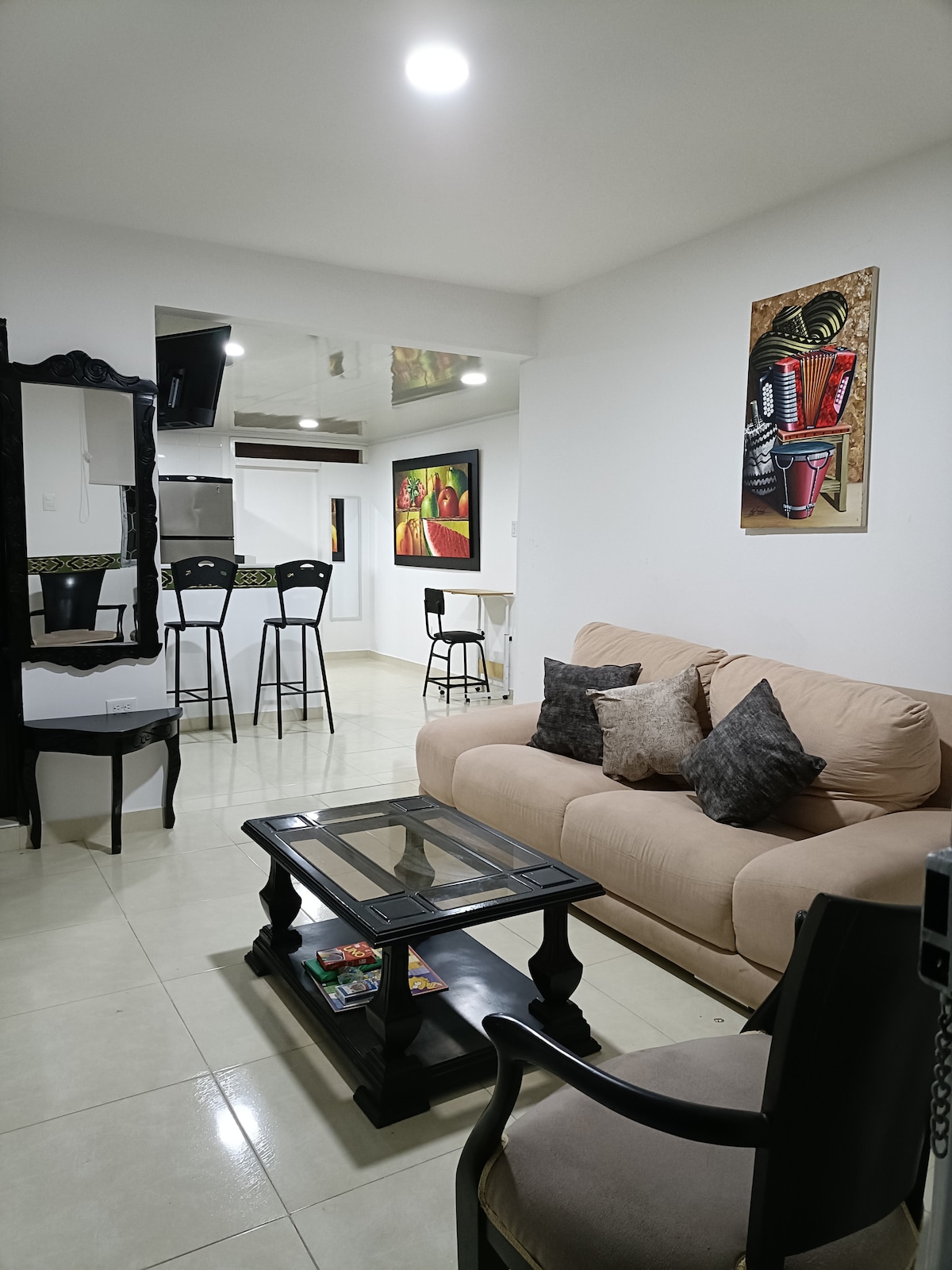 A living area features a beige sofa with decorative cushions, positioned near a dark coffee table. Two high-backed chairs complement the space, alongside a set of bar stools in the background. Artwork adorns the walls, enhancing the contemporary decor.