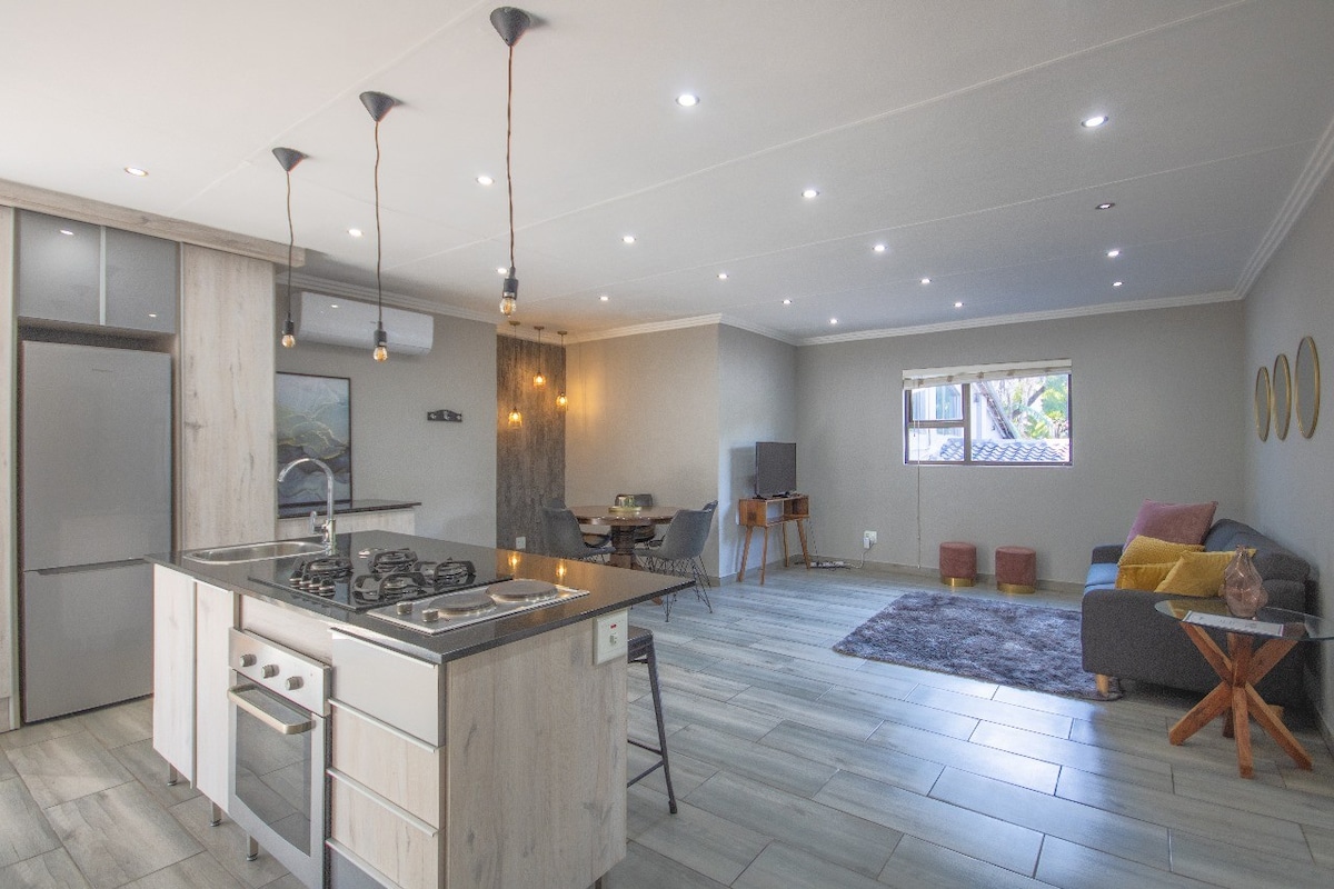 The open-plan kitchen and living area is illuminated by pendant lights, showcasing a modern kitchen island featuring a stovetop. A dining table for four is situated nearby, while a cozy lounge area is defined by a soft rug and adorned with contemporary furniture.