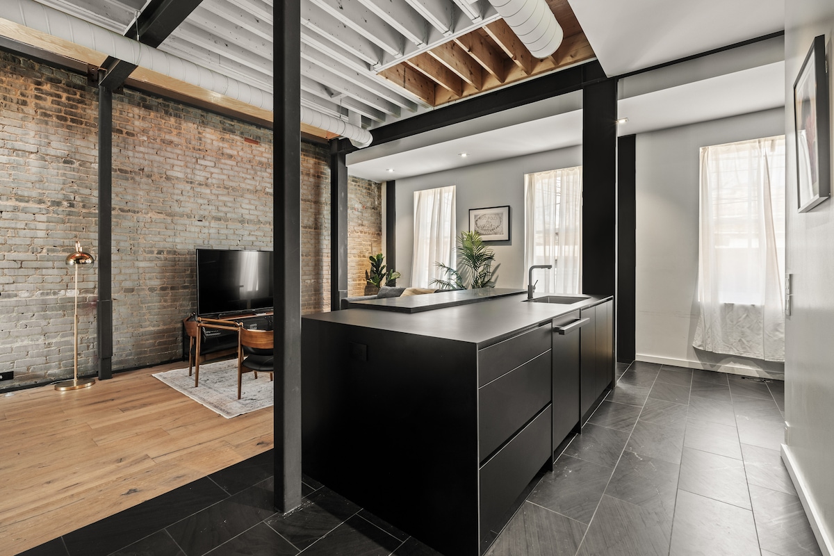 An open-concept kitchen features a modern black slate island, complemented by sleek cabinetry. Exposed brick walls add character, while a large window allows natural light to fill the space. A cozy living area with a TV and plants creates a welcoming atmosphere.